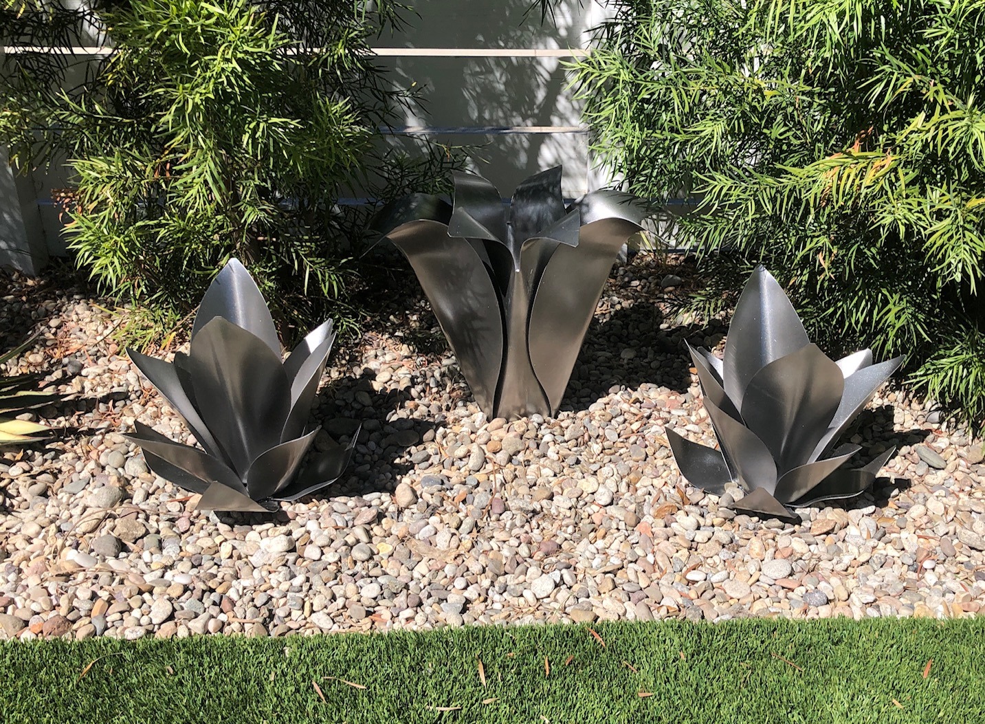 Three silver steel flower sculptures grouped in gravel beneath green planting